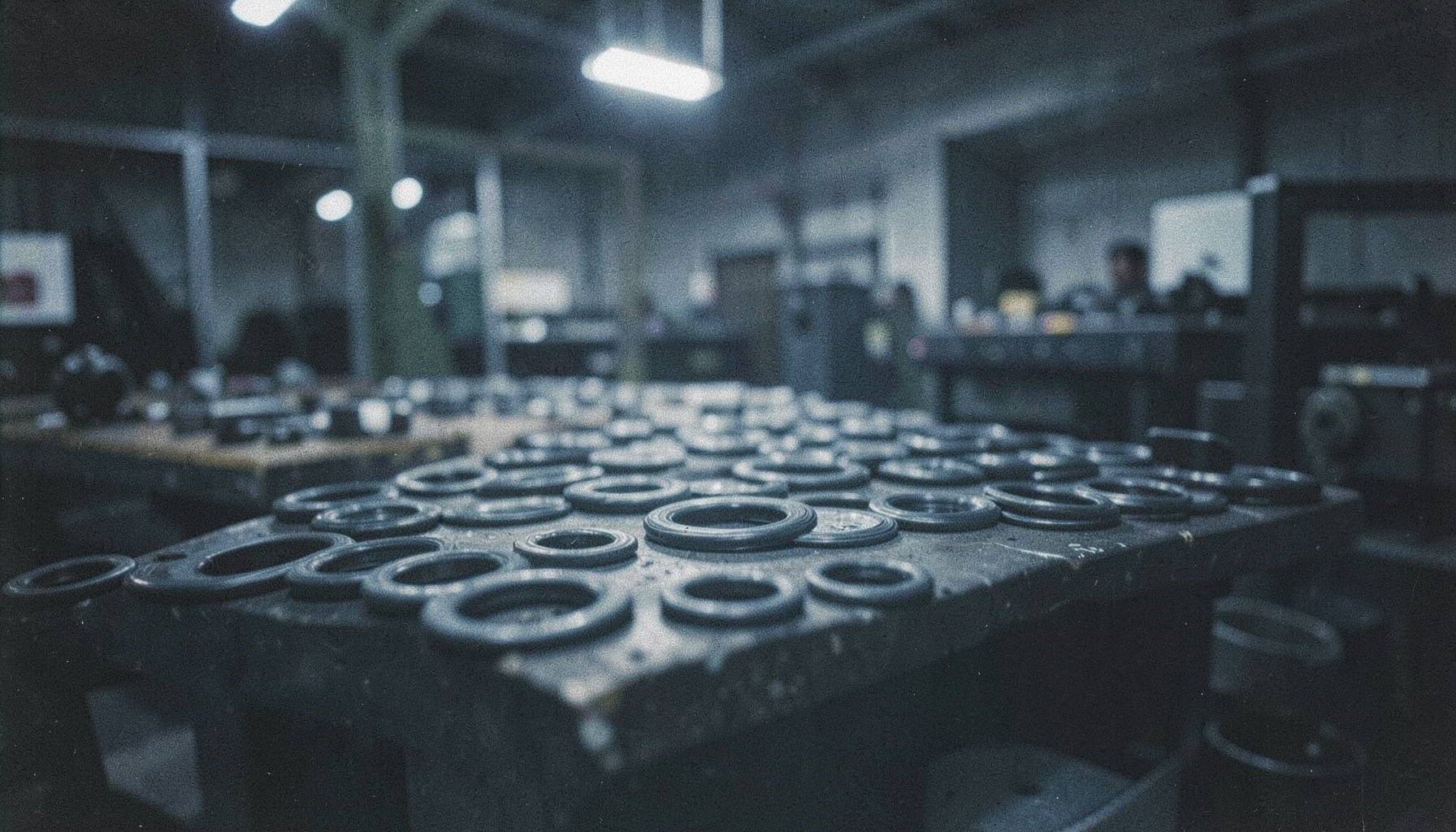 Rows of rubber oil seals on a factory workbench, dark industrial setting with soft overhead lighting, muted grays and blacks, film grain texture