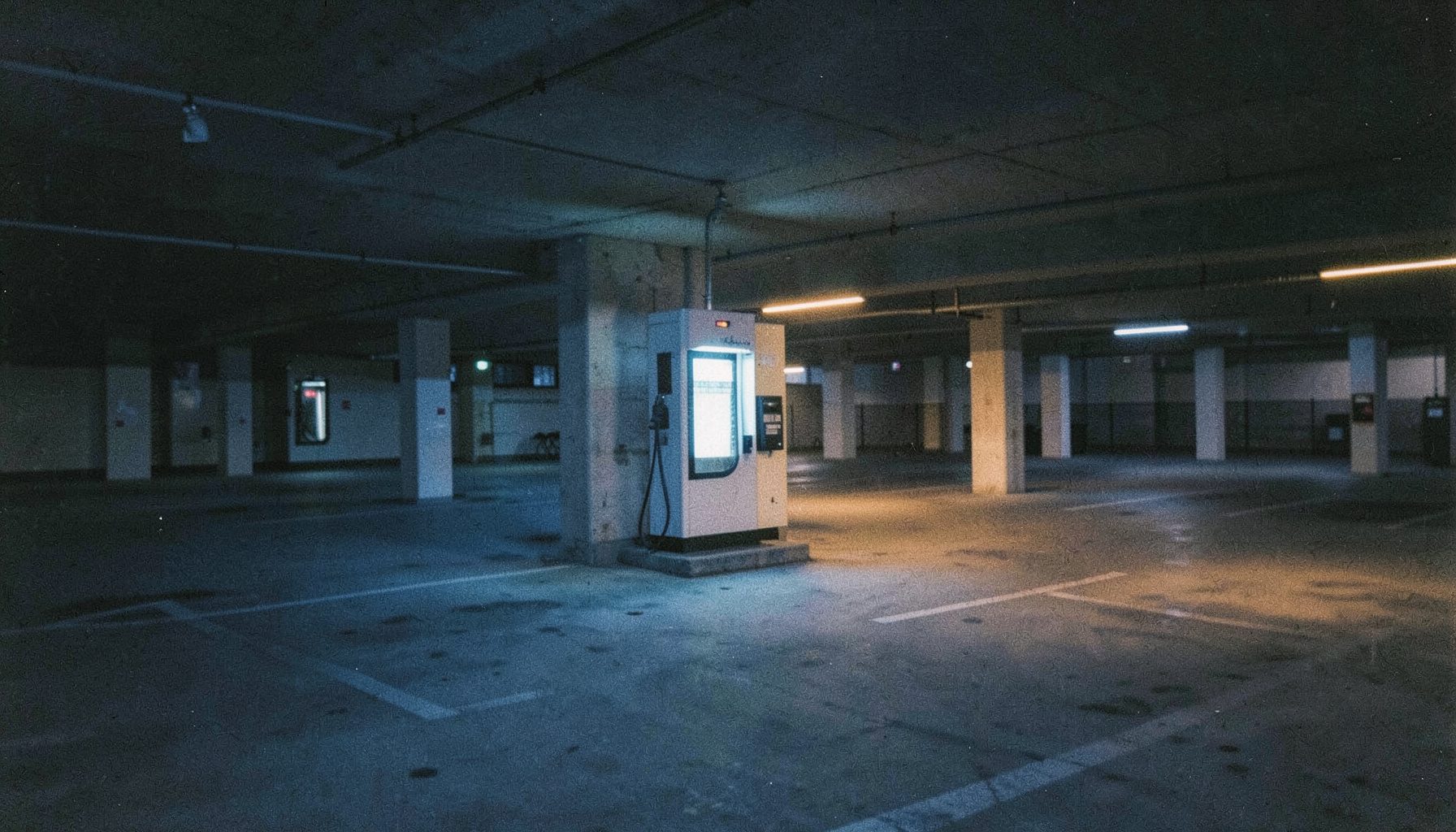 A solitary electric vehicle charging station glowing softly at twilight in an empty urban parking structure, cool blue and warm amber light, cinematic grain