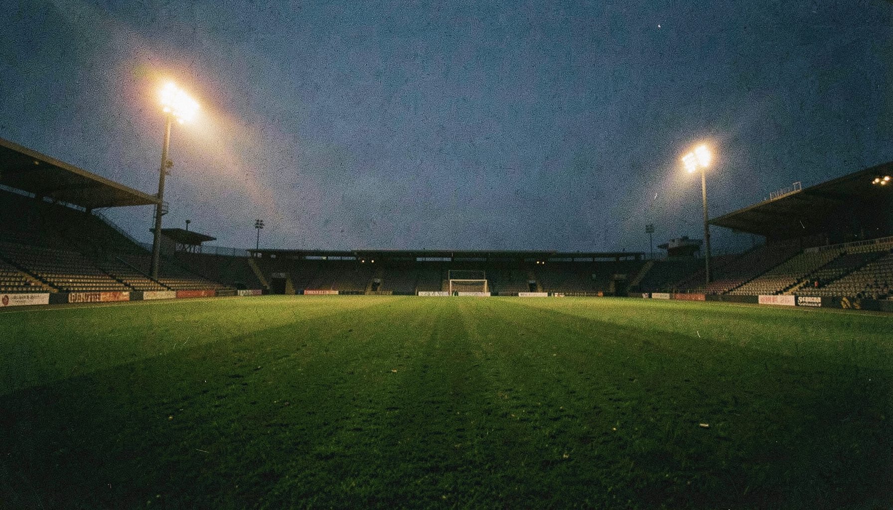 Wide-angle photograph of a floodlit football pitch at dusk, deep green turf under warm golden floodlights, empty stands in shadow, grainy film texture