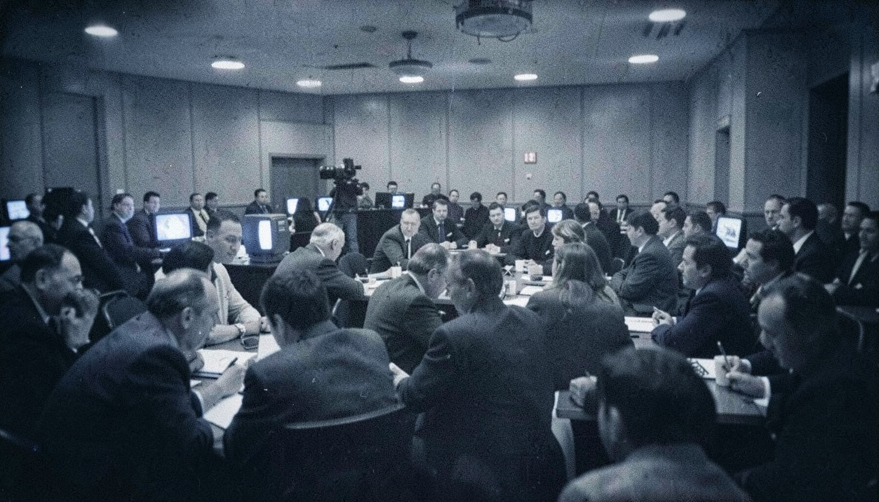 Grainy black-and-white film photograph of a crowded sports press room, journalists at desks, television monitors glowing softly