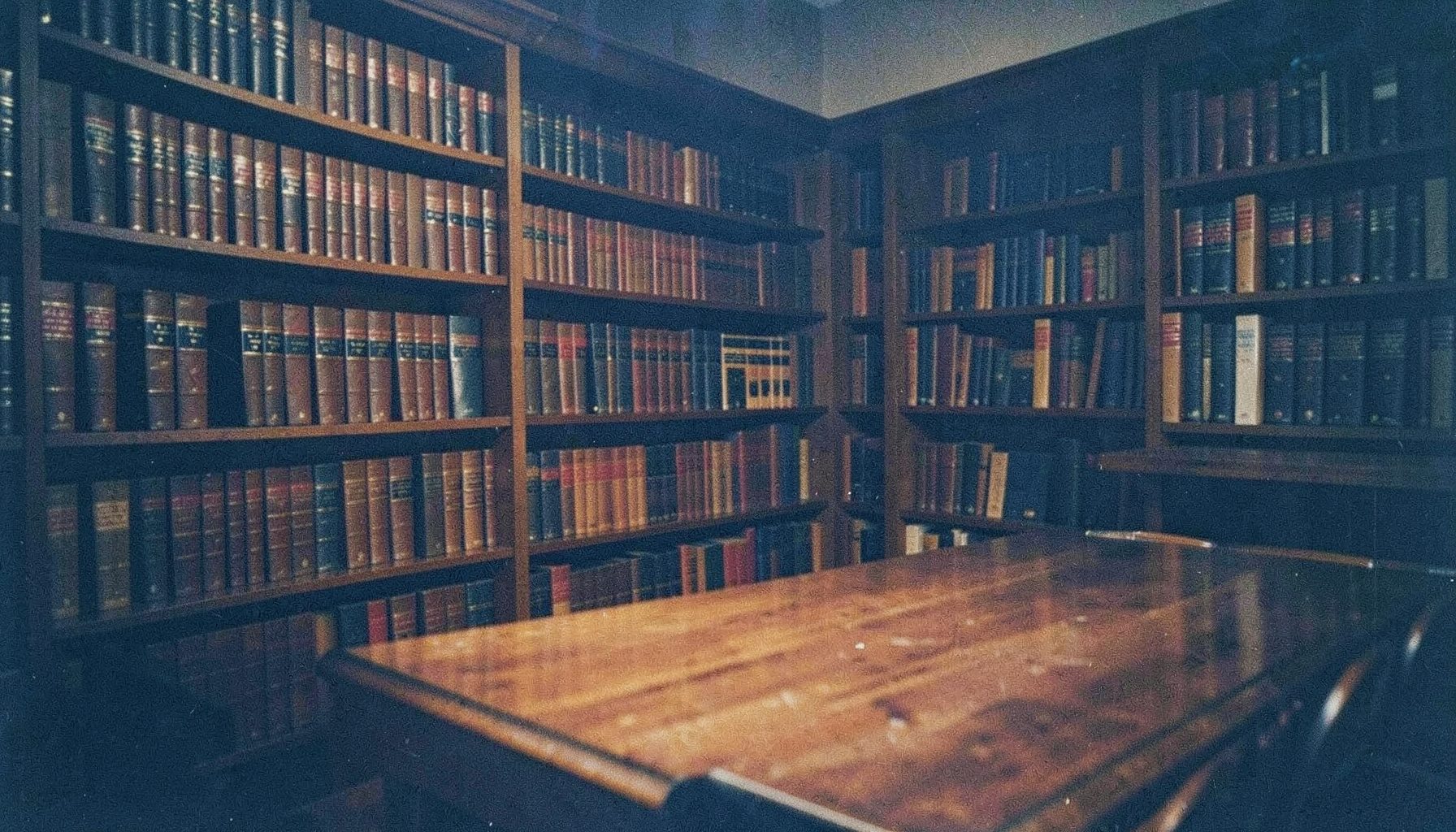 A dimly lit law library with rows of leather-bound books in deep brown and navy tones, warm amber light spilling across a polished wooden table, analog film grain visible