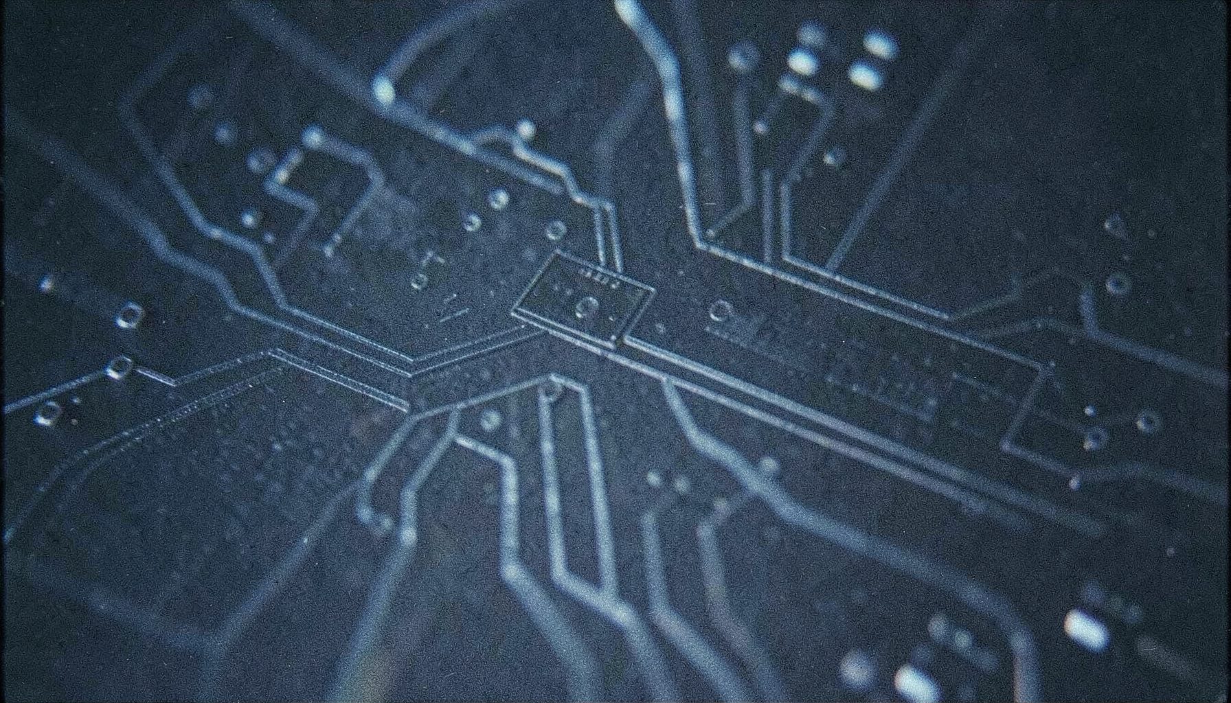 Abstract close-up of a dark silicon wafer surface with fine geometric circuit traces catching cool blue and silver reflections, grainy analog film texture