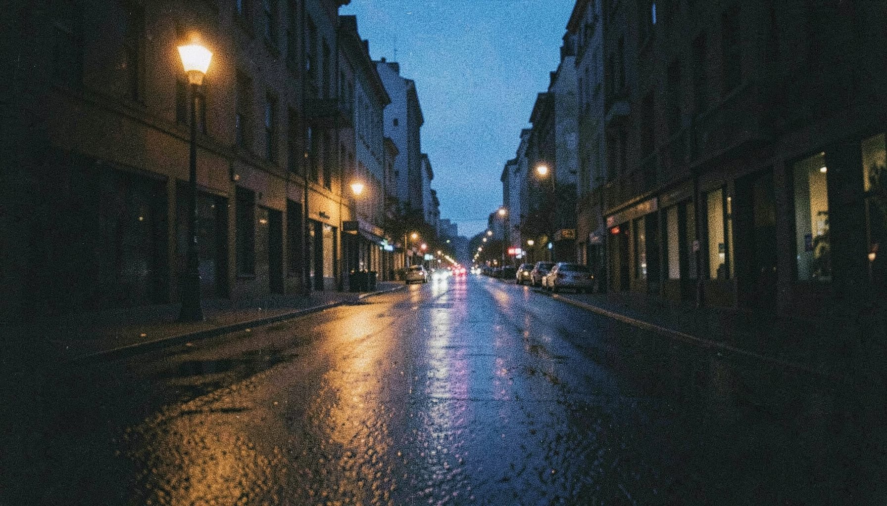A grainy analog photograph of a quiet city street at dusk, warm amber streetlights reflecting off wet pavement, deep navy shadows stretching across the frame, moody and contemplative atmosphere.