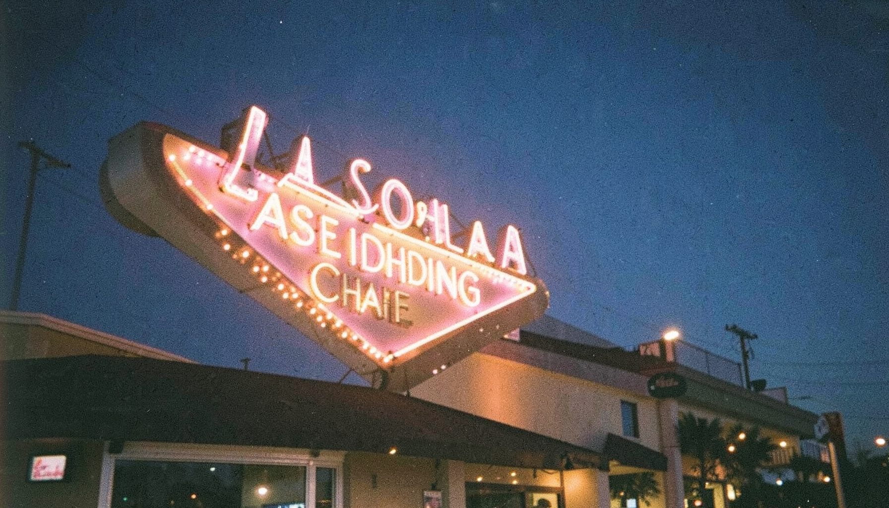 Neon-lit Las Vegas wedding chapel sign at dusk, warm pink and gold glow against a deep indigo sky, grainy film photograph